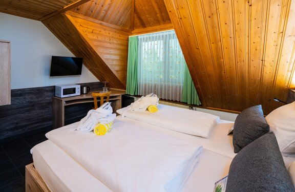Gemütliches Schlafzimmer mit Holzwänden, zwei Betten, TV, Mikrowelle und Fenster mit grünen Vorhängen